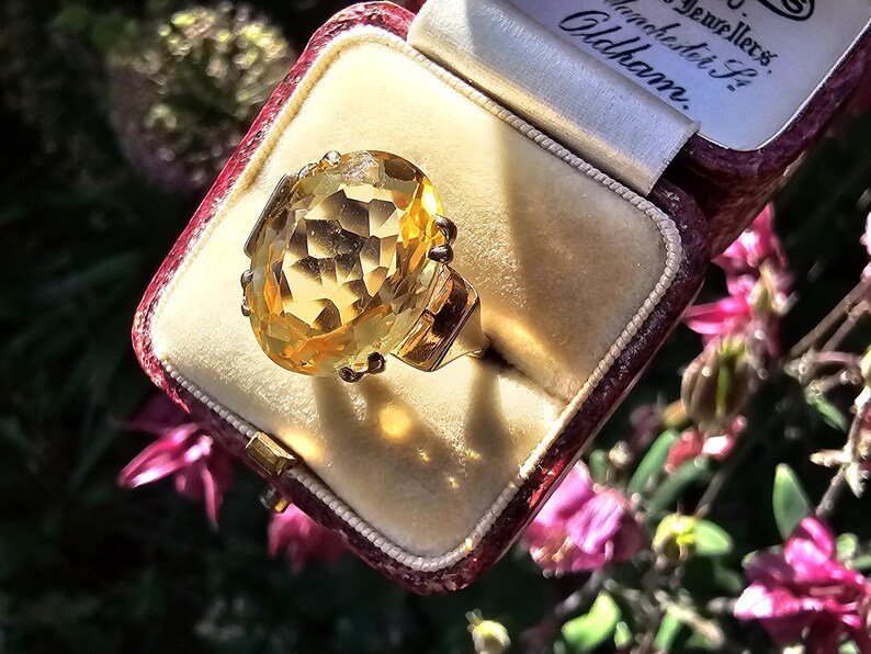 May include: A gold ring with a large, faceted, yellow gemstone, presented in a vintage maroon jewelry box with a cream-colored interior. The ring is the focal point, catching the light. The box is open, revealing the ring.