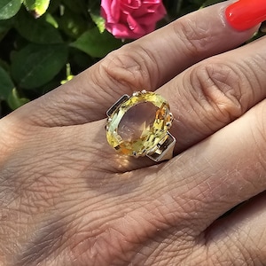 May include: A large, oval-cut yellow gemstone ring with a silver band. The ring's gemstone is the focal point, set in a silver band with decorative accents. The ring is on a hand with red nail polish, with a blurred background of green leaves and a pink flower.