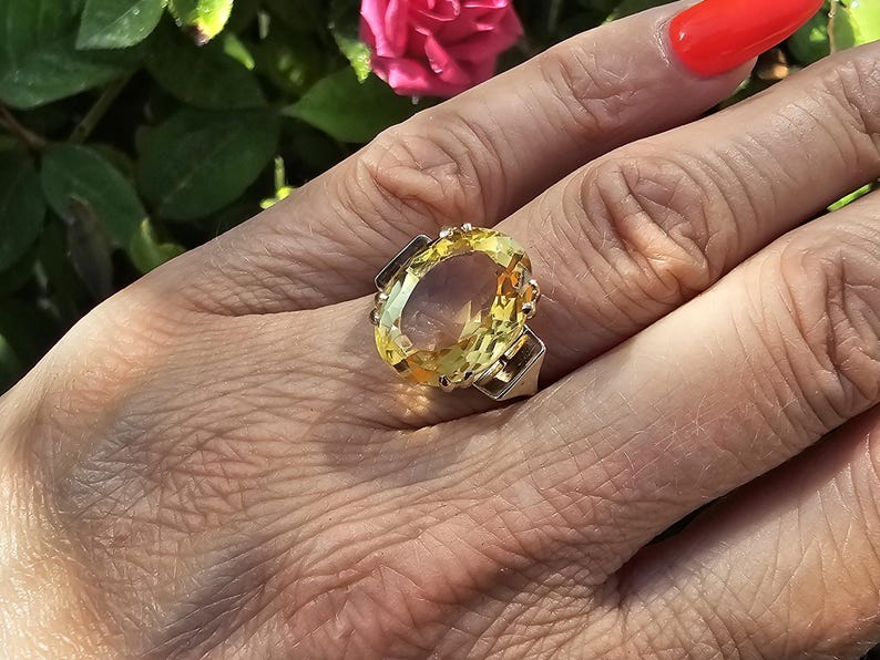 May include: A large, oval-cut yellow gemstone ring with a silver band. The ring's gemstone is the focal point, set in a silver band with decorative accents. The ring is on a hand with red nail polish, with a blurred background of green leaves and a pink flower.