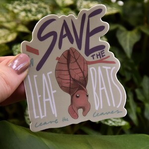 Op de afbeelding: Een beige sticker met de tekst "Save the Leaf Bats" in witte en paarse letters. De sticker bevat een cartoonillustratie van een bruine bladvleermuis met grote, afgeronde oren. De tekst "Leave the leaves" is ook in witte letters opgenomen.