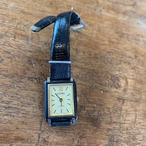 May include: A vintage rectangular FUTURA quartz watch with a black leather band. The watch face is gold with black Roman numerals and hands. The watch band is black and shows signs of wear.