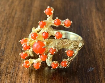 May include: A gold-toned ring featuring a coral-like design with numerous small, round, orange gemstones. The ring has a textured band and a cluster of the orange stones, creating a unique and eye-catching piece of jewelry.