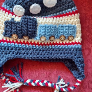 May include: A blue and white crocheted hat with a train design. The hat has a white pom pom on top and a long, braided cord with red, white, and blue yarn.