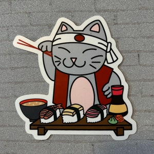 May include: A grey cat wearing a red and white headband holds chopsticks and looks at a plate of sushi. The plate has four pieces of sushi, a small bowl of soup, a bottle of soy sauce, and a small green piece of wasabi.