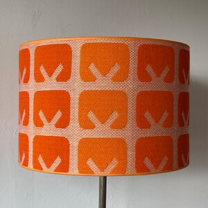 Op de afbeelding: Oranje en witte lampenkap met geometrisch patroon en een metalen voet.