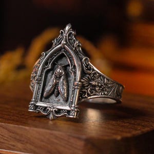 Può includere: Un anello in metallo color argento caratterizzato da un intricato design ad arco con un rilievo di falena al centro. La fascia presenta incisioni dettagliate sui lati. Un'etichetta fissata alla base riporta il testo GR-001 e Acherontia atropos.