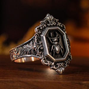 Può includere: Un anello color argento con una faccia ottagonale caratterizzata da un rilievo di cervo volante. La fascia e l'incastonatura sono decorate con complessi motivi a volute e incisioni a forma di foglia. L'anello è posizionato su una scatola di legno etichettata GR-002.