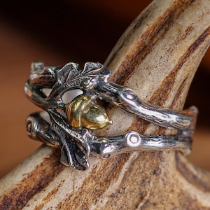 Puede incluir: Un conjunto de anillos plateados con un diseño inspirado en la naturaleza. El anillo tiene una banda en forma de rama con detalles de hojas y bellotas. La bellota es de color dorado. El anillo se muestra sobre una cornamenta.