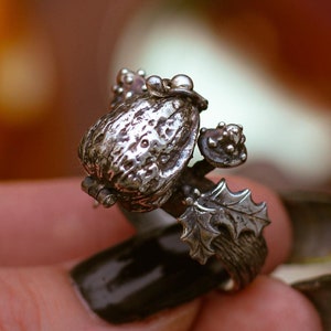 Può includere: Un anello d'argento con un design di ghianda dettagliato. La ghianda è circondata da foglie e bacche.