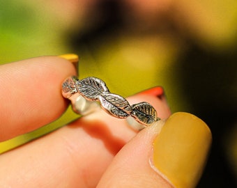 May include: A silver ring with a leaf design. The ring is being held by a hand with yellow nail polish.