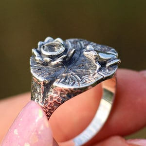 May include: A silver ring featuring a detailed design of a frog sitting on a lily pad with a flower. The ring is worn on a finger with pink nail polish.