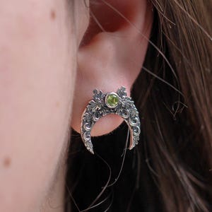 May include: A silver crescent moon earring with intricate detailing and a small green gemstone at the center. The earring is worn on an ear, showcasing its design and craftsmanship. The jewelry is a stud earring.