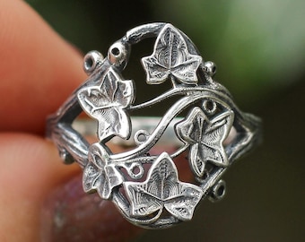 May include: Silver-toned ring featuring an intricate ivy leaf design. The ring showcases detailed leaves and vines, creating a nature-inspired aesthetic. The ring is held between fingers, with a blurred green background.