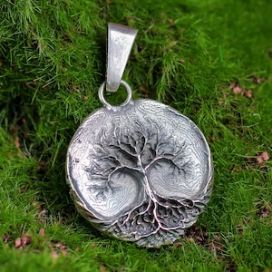 May include: A silver pendant with a detailed tree of life design. The pendant is round with a textured surface, and hangs from a silver bail. The pendant is set against a backdrop of green moss.