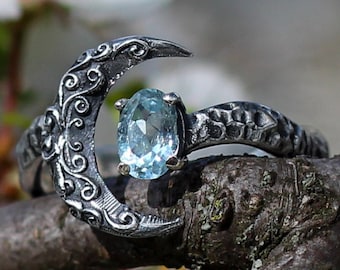 Puede incluir: Un anillo de plata con un diseño de luna creciente y una piedra preciosa azul claro. El anillo está colocado en una rama con flores blancas.