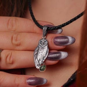 May include: A silver owl pendant with a green stone hangs from a black cord necklace. A silver ring with a blue stone is on a finger. The owl pendant is detailed with etched feathers and a textured surface.