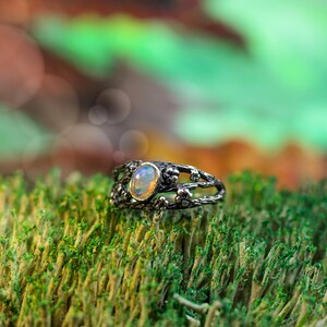 May include: A silver ring with a delicate, branch-like design and a white opal gemstone set in the center. The ring is resting on a bed of green moss.