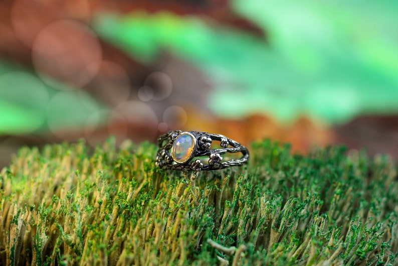 May include: A silver ring with a delicate, branch-like design and a white opal gemstone set in the center. The ring is resting on a bed of green moss.