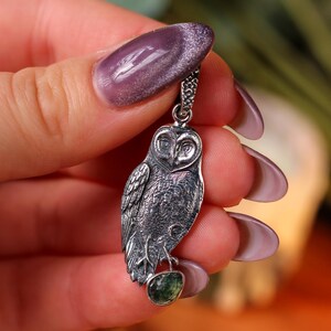 May include: A detailed silver owl pendant, with a small green gemstone at the base. The owl is suspended from a fine chain. The pendant is held up against a blurred background.