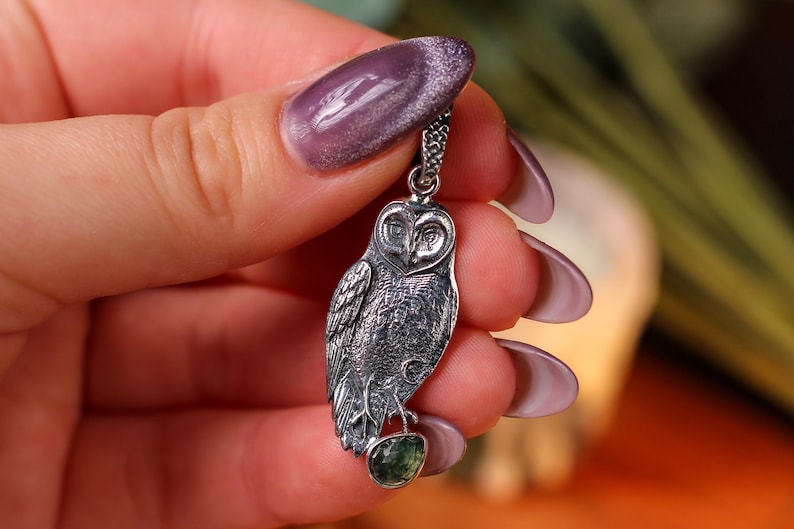 May include: A detailed silver owl pendant, with a small green gemstone at the base. The owl is suspended from a fine chain. The pendant is held up against a blurred background.