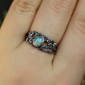 May include: A silver ring with a delicate, intricate design featuring a branch-like pattern and a white opal gemstone.