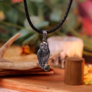 May include: A silver owl pendant suspended from a black, plaited cord. The owl features intricate detailing and a green gemstone. The background includes a wooden antler, a candle, and foliage, evoking a natural theme.