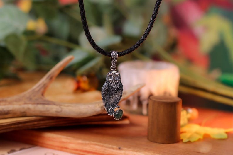 May include: A silver owl pendant suspended from a black, plaited cord. The owl features intricate detailing and a green gemstone. The background includes a wooden antler, a candle, and foliage, evoking a natural theme.