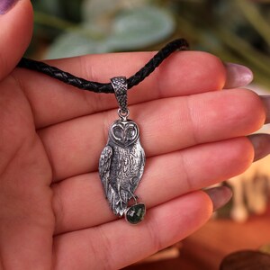 May include: Silver owl pendant on a black cord. The owl pendant has detailed feathering and a small green gemstone. The pendant is held in a hand with purple nail polish.