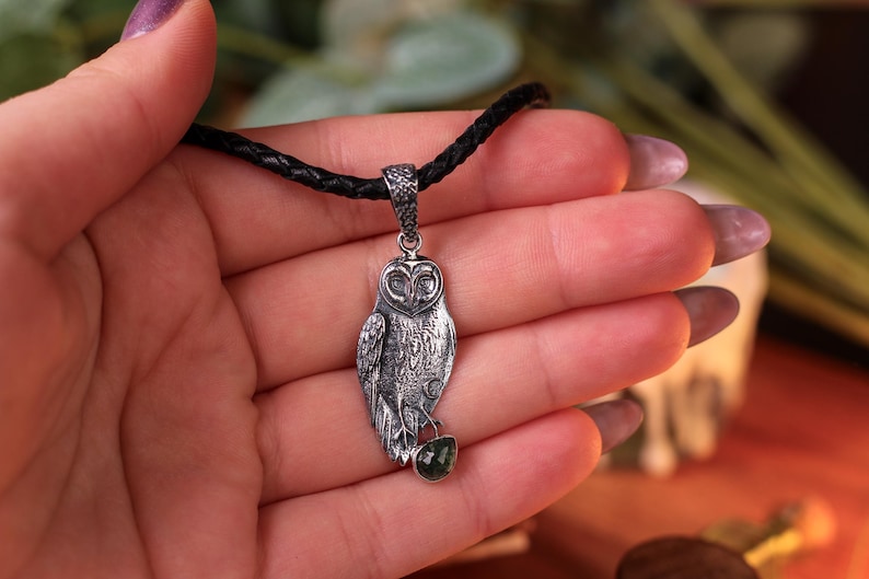 May include: Silver owl pendant on a black cord. The owl pendant has detailed feathering and a small green gemstone. The pendant is held in a hand with purple nail polish.