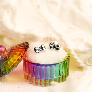 May include: A pair of silver bow-shaped earrings in a rainbow-colored glass jewelry box with a white velvet lining.