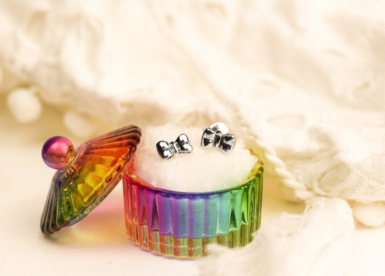 May include: A pair of silver bow-shaped earrings in a rainbow-colored glass jewelry box with a white velvet lining.