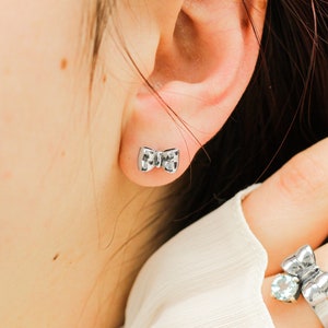 May include: A pair of silver bow-shaped earrings. The earrings are small and delicate, and they are a popular style of jewelry.