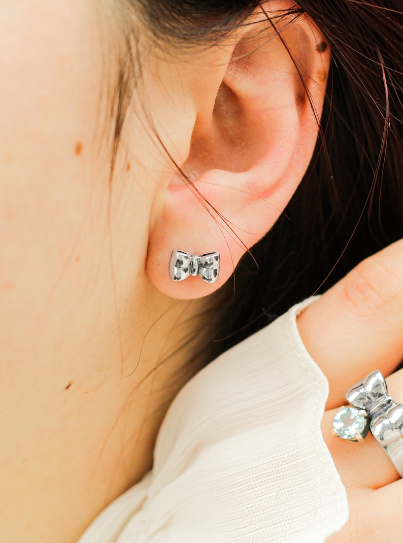 May include: A pair of silver bow-shaped earrings. The earrings are small and delicate, and they are a popular style of jewelry.