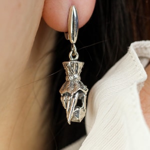 May include: A silver earring featuring a detailed hand motif holding a faceted gem. The earring hangs from a simple, oval clasp. The design has a textured, antique appearance, with a gothic style.