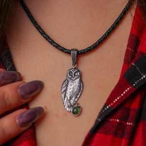 May include: A silver owl pendant necklace with a black braided cord. The owl pendant has detailed feathering and a small green gemstone. The necklace is displayed against a red and black plaid shirt.