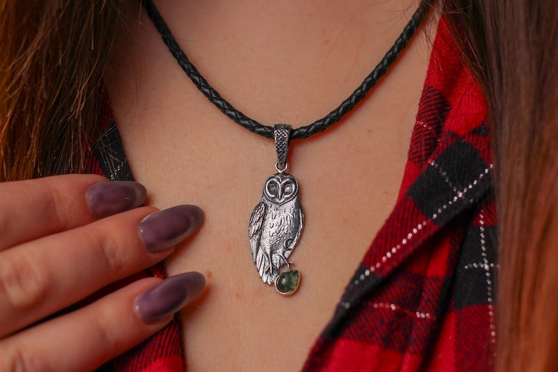 May include: A silver owl pendant necklace with a black braided cord. The owl pendant has detailed feathering and a small green gemstone. The necklace is displayed against a red and black plaid shirt.