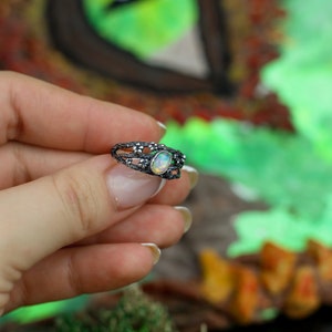 May include: A silver ring with a delicate, intricate design featuring a white opal gemstone. The ring is set against a blurred background of green and brown.