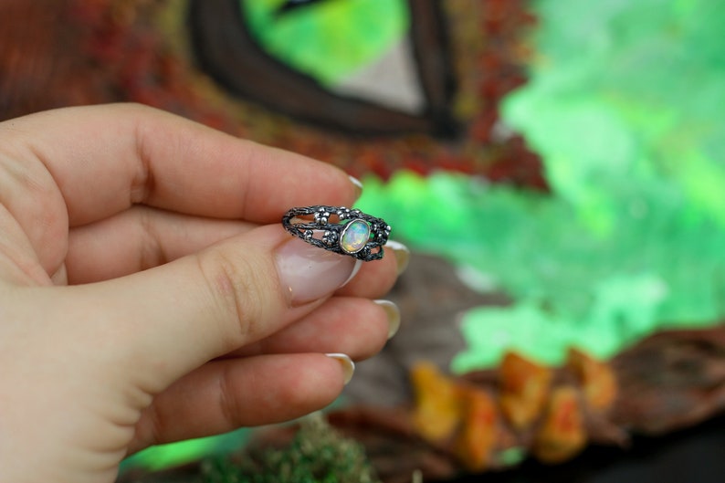 May include: A silver ring with a delicate, intricate design featuring a white opal gemstone. The ring is set against a blurred background of green and brown.