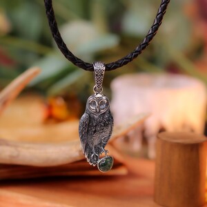 May include: A silver owl pendant on a black braided cord. The owl has detailed feathering and is perched on a green stone. The pendant hangs against a blurred background with a wooden element and greenery.