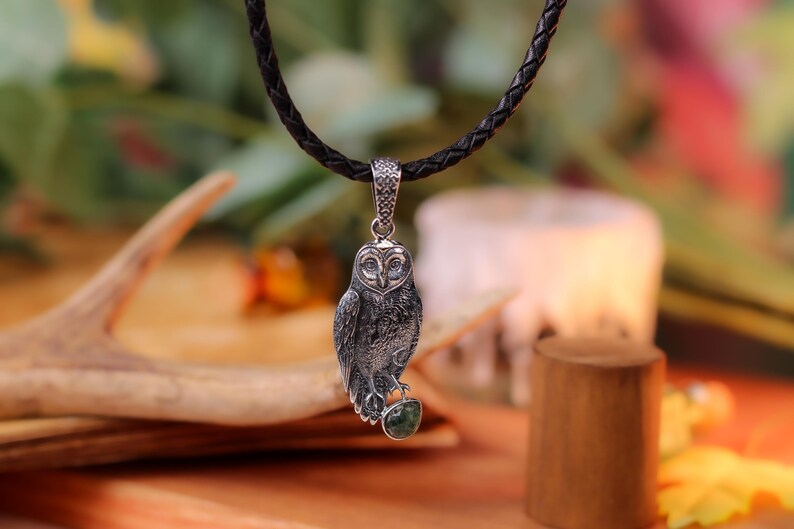 May include: A silver owl pendant on a black braided cord. The owl has detailed feathering and is perched on a green stone. The pendant hangs against a blurred background with a wooden element and greenery.