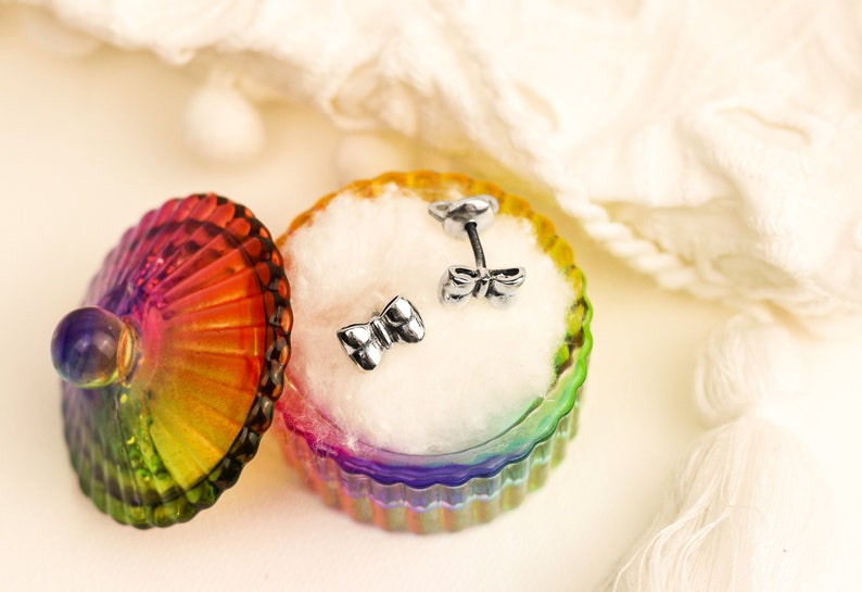 May include: A pair of silver bow-shaped earrings in a rainbow-colored glass jewelry box with a white cotton lining.