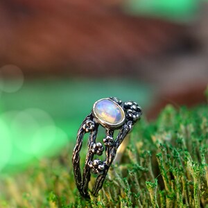 May include: A silver ring with a delicate floral design and an oval opal gemstone. The ring is set on a bed of green moss.