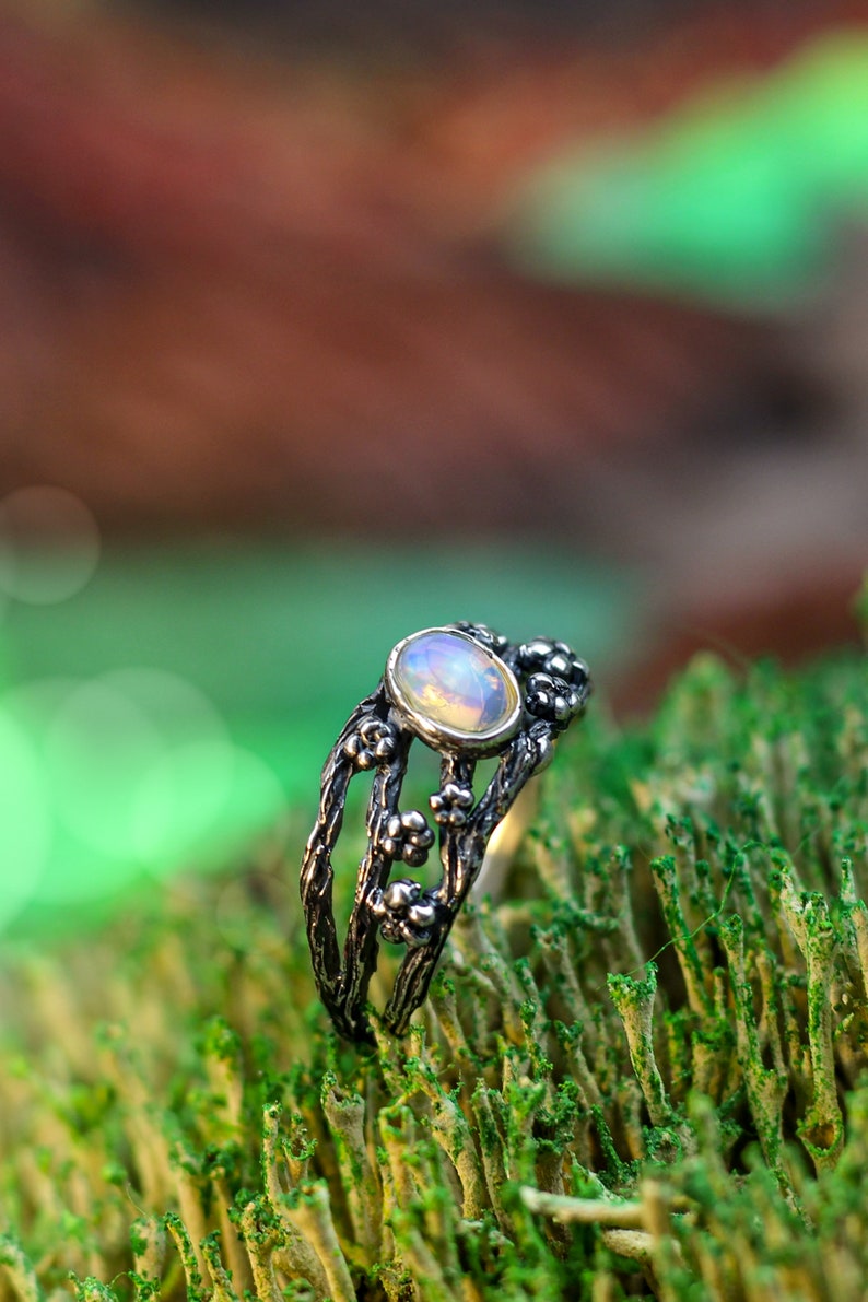 May include: A silver ring with a delicate floral design and an oval opal gemstone. The ring is set on a bed of green moss.