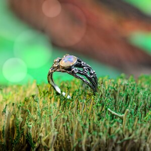 May include: A silver ring with a white opal gemstone set in a delicate, twig-like design. The ring is resting on a bed of green moss.