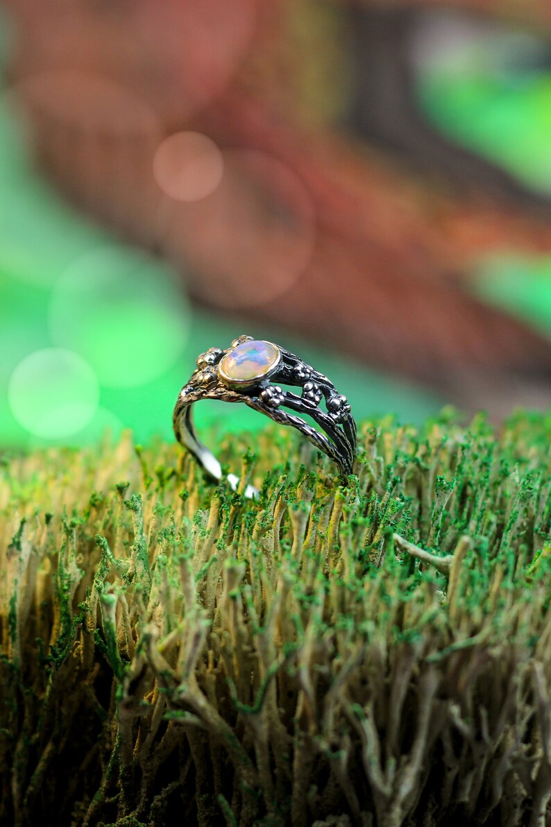 May include: A silver ring with a white opal gemstone set in a delicate, twig-like design. The ring is resting on a bed of green moss.