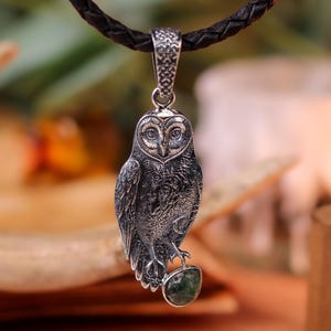 May include: A silver owl pendant on a dark brown leather cord. The owl pendant features detailed craftsmanship and holds a green stone. The pendant hangs from a braided leather cord. The background is blurred, with warm tones.