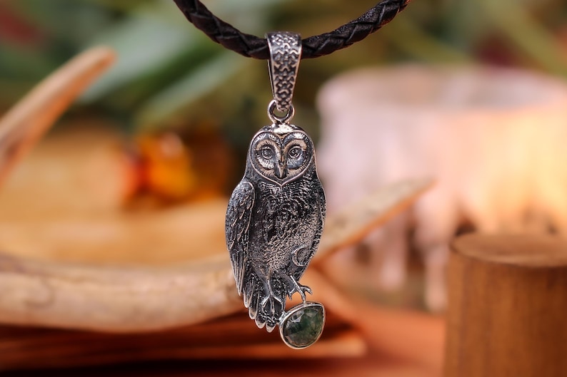May include: A silver owl pendant on a dark brown leather cord. The owl pendant features detailed craftsmanship and holds a green stone. The pendant hangs from a braided leather cord. The background is blurred, with warm tones.