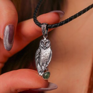 May include: Silver owl pendant necklace with a black cord. The owl pendant has detailed feathering and holds a small green gemstone. The necklace is held by a person with purple nail varnish.