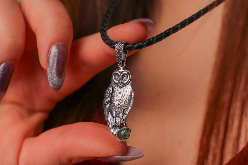 May include: Silver owl pendant necklace with a black cord. The owl pendant has detailed feathering and holds a small green gemstone. The necklace is held by a person with purple nail varnish.
