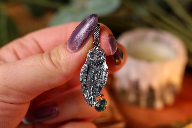 May include: A detailed silver owl pendant with a dark patina, featuring intricate feather detailing and a small green gemstone. The owl is suspended from a delicate chain, showcasing a gothic or fantasy theme.